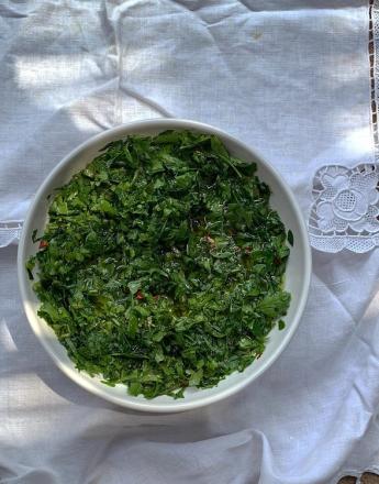 A white bowl of vibrant green chopped herbs with olive oil and red flecks on a white lace tablecloth.