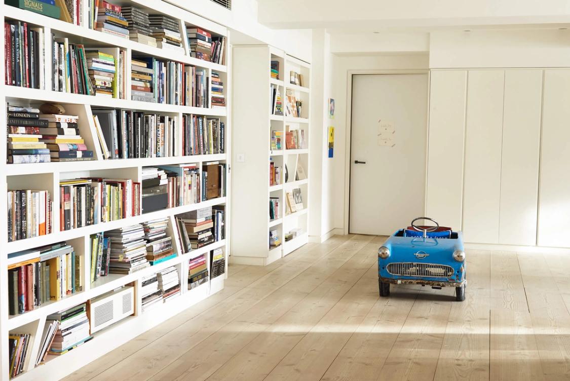 A bright room with white bookshelves packed with books, a light wooden floor, and a blue vintage pedal car.