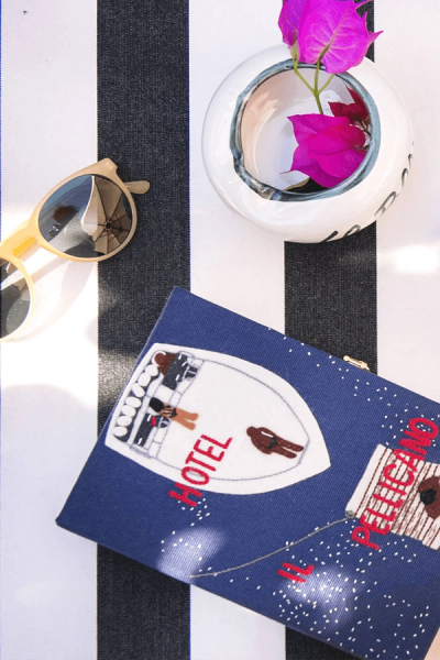A blue embroidered clutch with "Hotel Il Pelicano," yellow sunglasses reflecting an umbrella, and a white vase with pink flowers on a black and white striped surface.