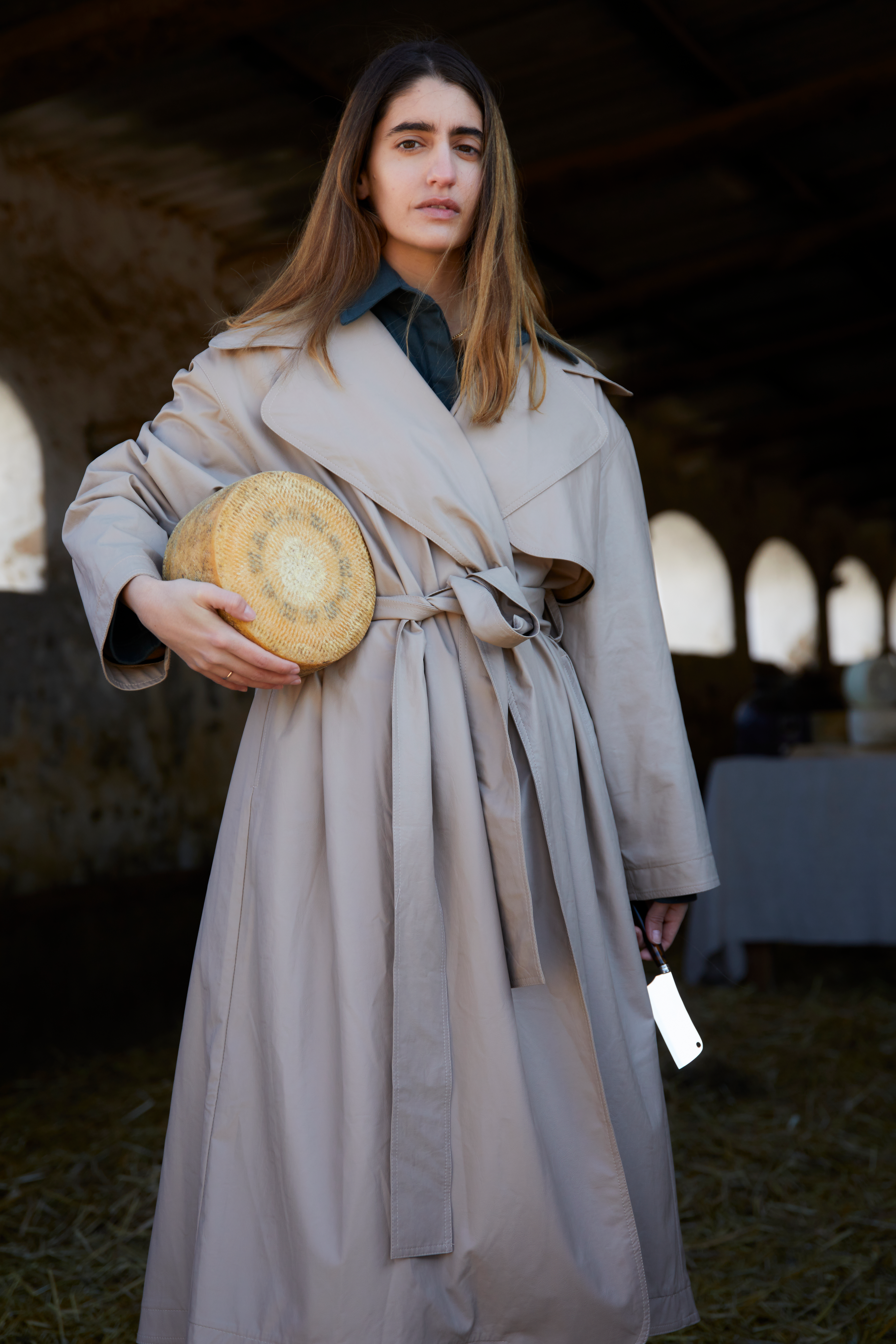 A woman in a beige trench coat holds a round cheese and a cleaver, standing in a rustic barn.