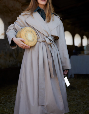 A woman in a beige trench coat holds a round cheese and a cleaver, standing in a rustic barn.