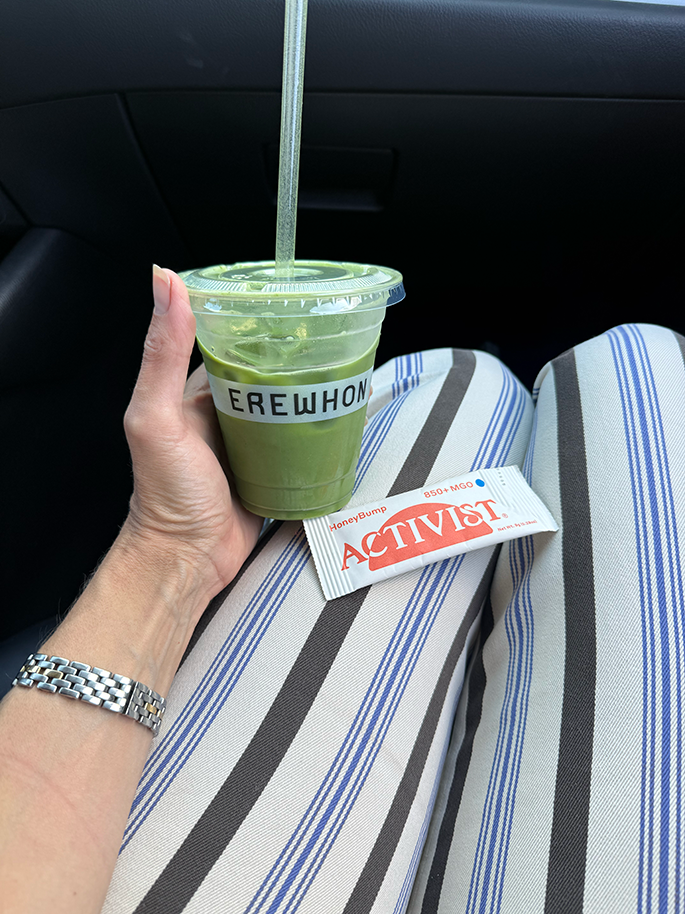 A hand holds an Erewhon green drink with a straw, an Activist packet rests on striped pants, inside a car.