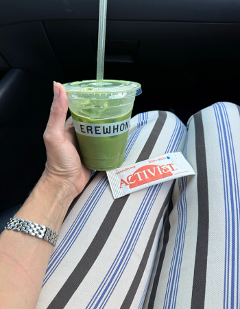 A hand holds an Erewhon green drink with a straw, an Activist packet rests on striped pants, inside a car.