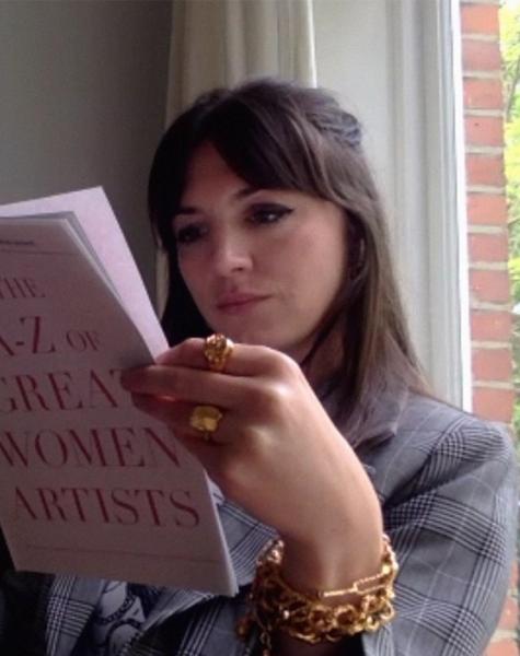 A woman with dark hair and gold jewelry reads "The A-Z of Great Women Artists" book.