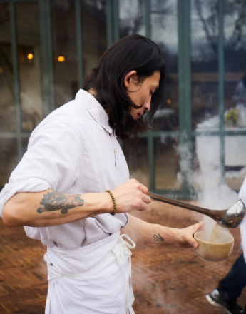 A person with long dark hair and tattooed arms pours steaming liquid from a ladle into a bowl.