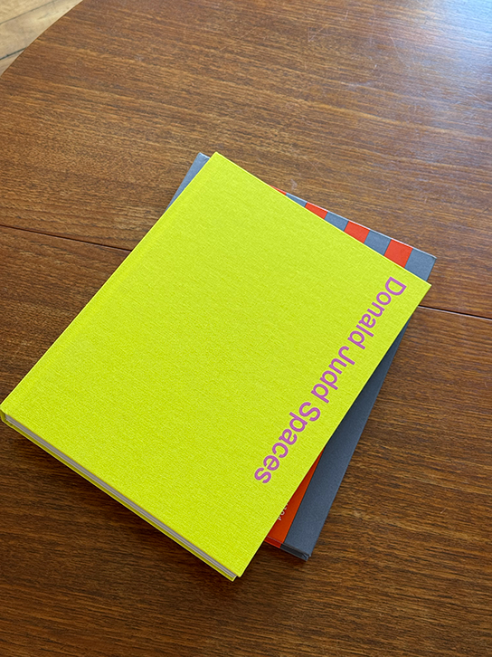 A bright lime green book titled 'Donald Judd Spaces' is stacked on top of a dark gray book on a wooden table.