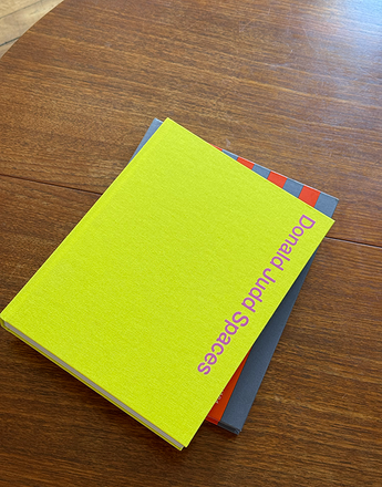 A bright lime green book titled 'Donald Judd Spaces' is stacked on top of a dark gray book on a wooden table.