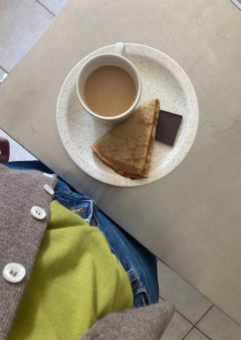 A white speckled plate on a light table holds a cup of coffee, a folded crepe, and a dark chocolate square, with a person's lap in a gray cardigan and jeans visible in the foreground.