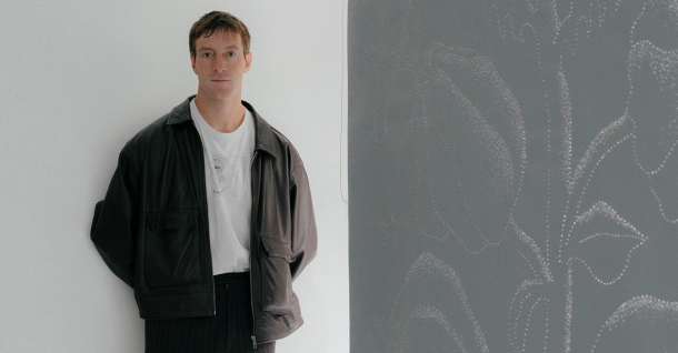 A man in a dark jacket stands beside a grey art panel with a white dotted floral design.
