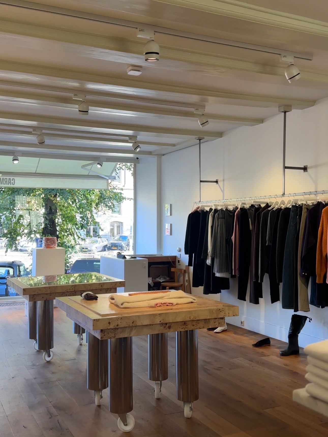Interior of a modern clothing boutique with wooden display tables, a long clothing rack, and a large window looking out onto a street.