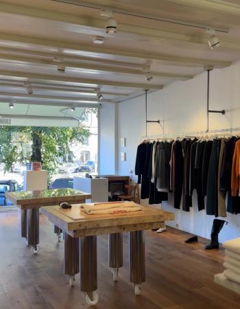 Interior of a modern clothing boutique with wooden display tables, a long clothing rack, and a large window looking out onto a street.