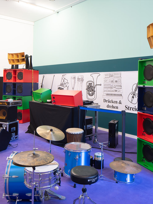A blue drum kit sits on a purple carpet, surrounded by tall stacks of colorful red, blue, and green speakers. The wall behind has musical instrument illustrations.