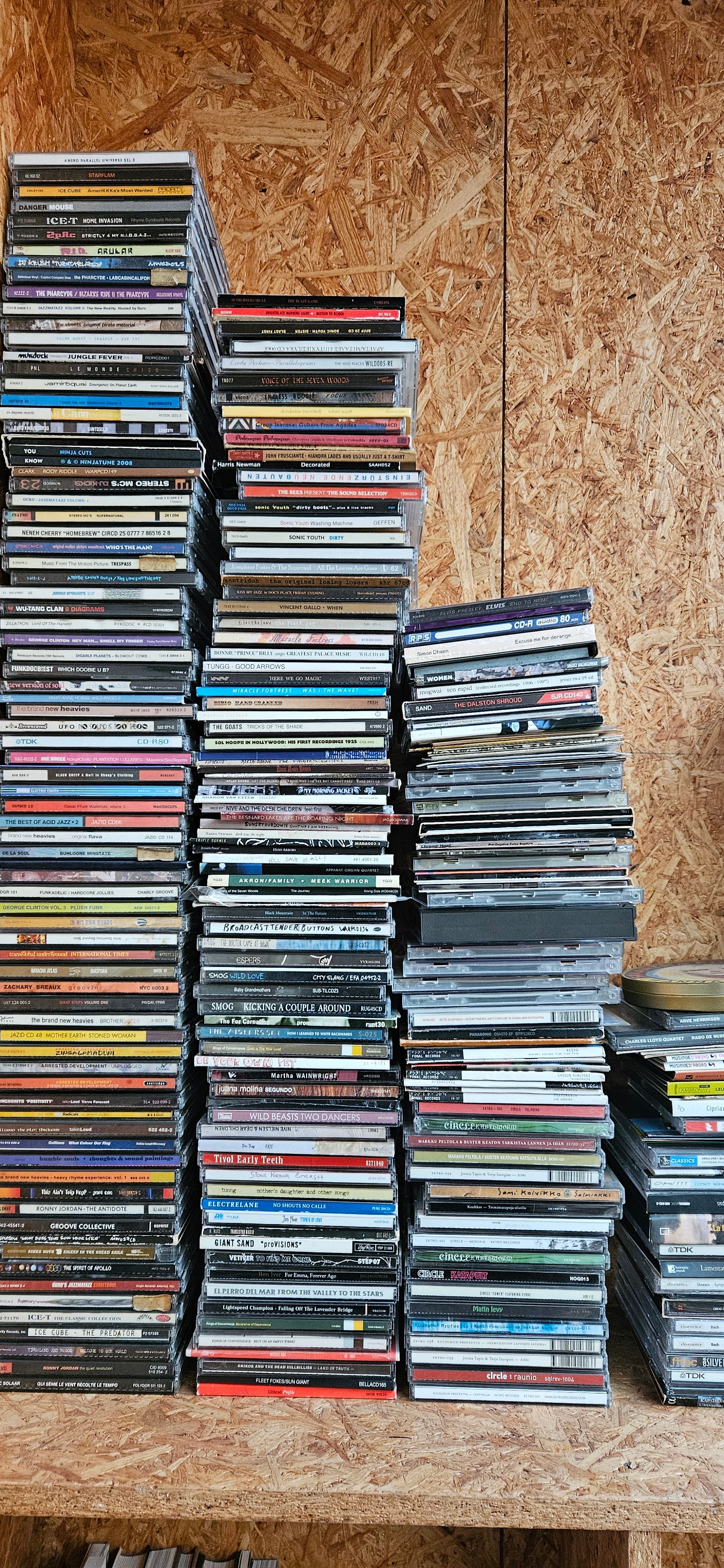 Tall stacks of CDs on a wooden shelf against an OSB wall.