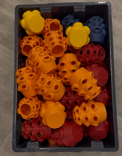 A dark crate filled with numerous colorful, bumpy construction toy pieces in orange, red, yellow, and blue.