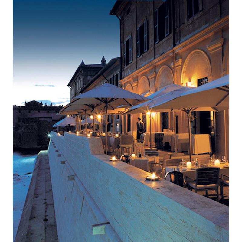 An illuminated riverside restaurant terrace at dusk, with umbrellas, tables, and candlelight.