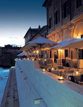An illuminated riverside restaurant terrace at dusk, with umbrellas, tables, and candlelight.