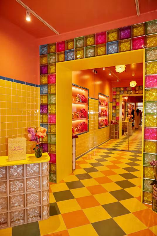 A vibrant store interior featuring yellow tiled walls, an orange ceiling, and a yellow archway framed by a wall of multicolored glass blocks. The floor has a checkered pattern, and a "Susan Alexandra" bag sits on a glass block counter.