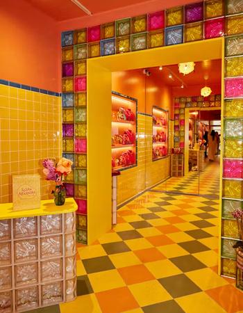 A vibrant store interior featuring yellow tiled walls, an orange ceiling, and a yellow archway framed by a wall of multicolored glass blocks. The floor has a checkered pattern, and a "Susan Alexandra" bag sits on a glass block counter.