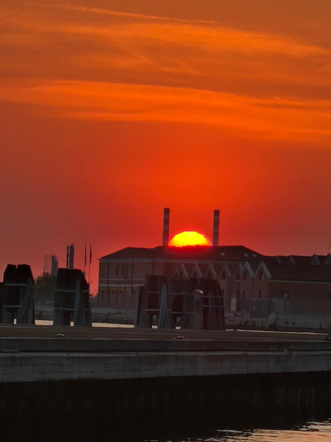 Vibrant red sunset with a yellow sun appearing between two smokestacks on a dark building, above a pier.