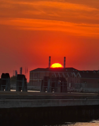 Vibrant red sunset with a yellow sun appearing between two smokestacks on a dark building, above a pier.