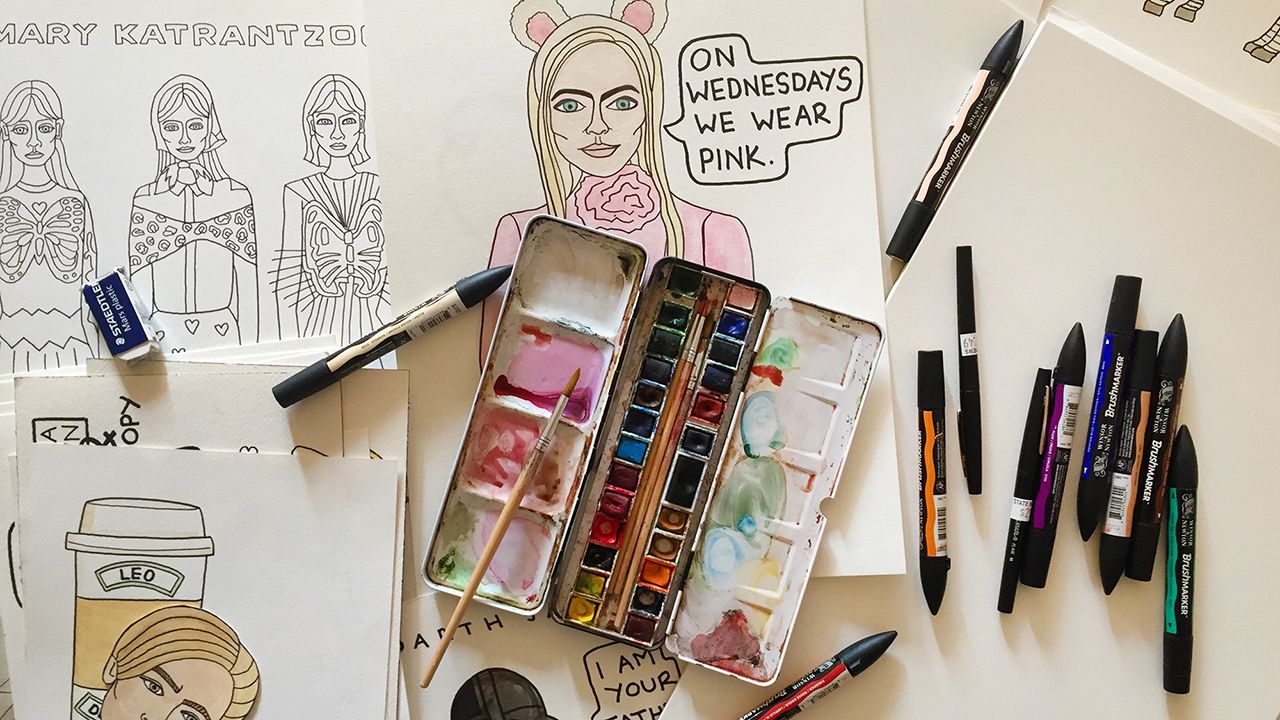 A watercolor palette, markers, and pencils are surrounded by hand-drawn sketches, including one of a woman with bunny ears and the quote "On Wednesdays we wear pink."