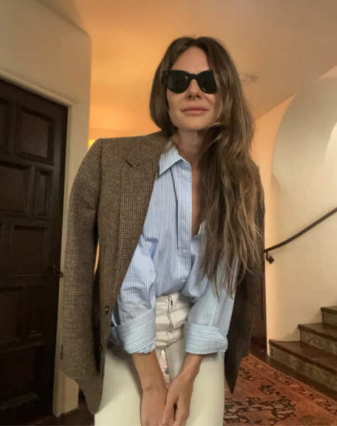A woman in sunglasses wears a brown plaid blazer draped over a blue striped shirt and light pants, standing indoors.