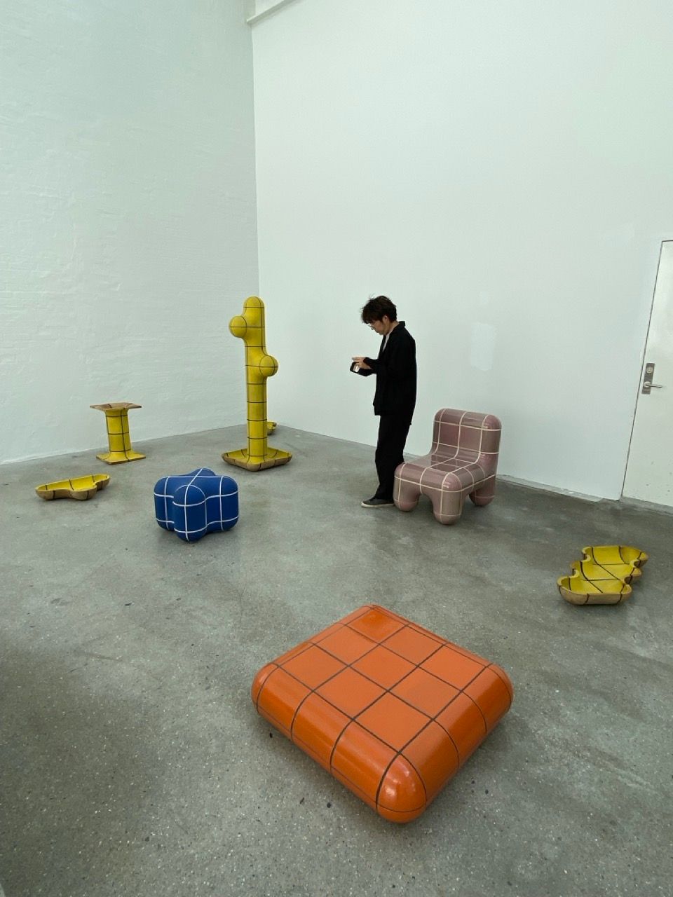 A person in a minimalist gallery observes colorful, grid-patterned art furniture and sculptures.