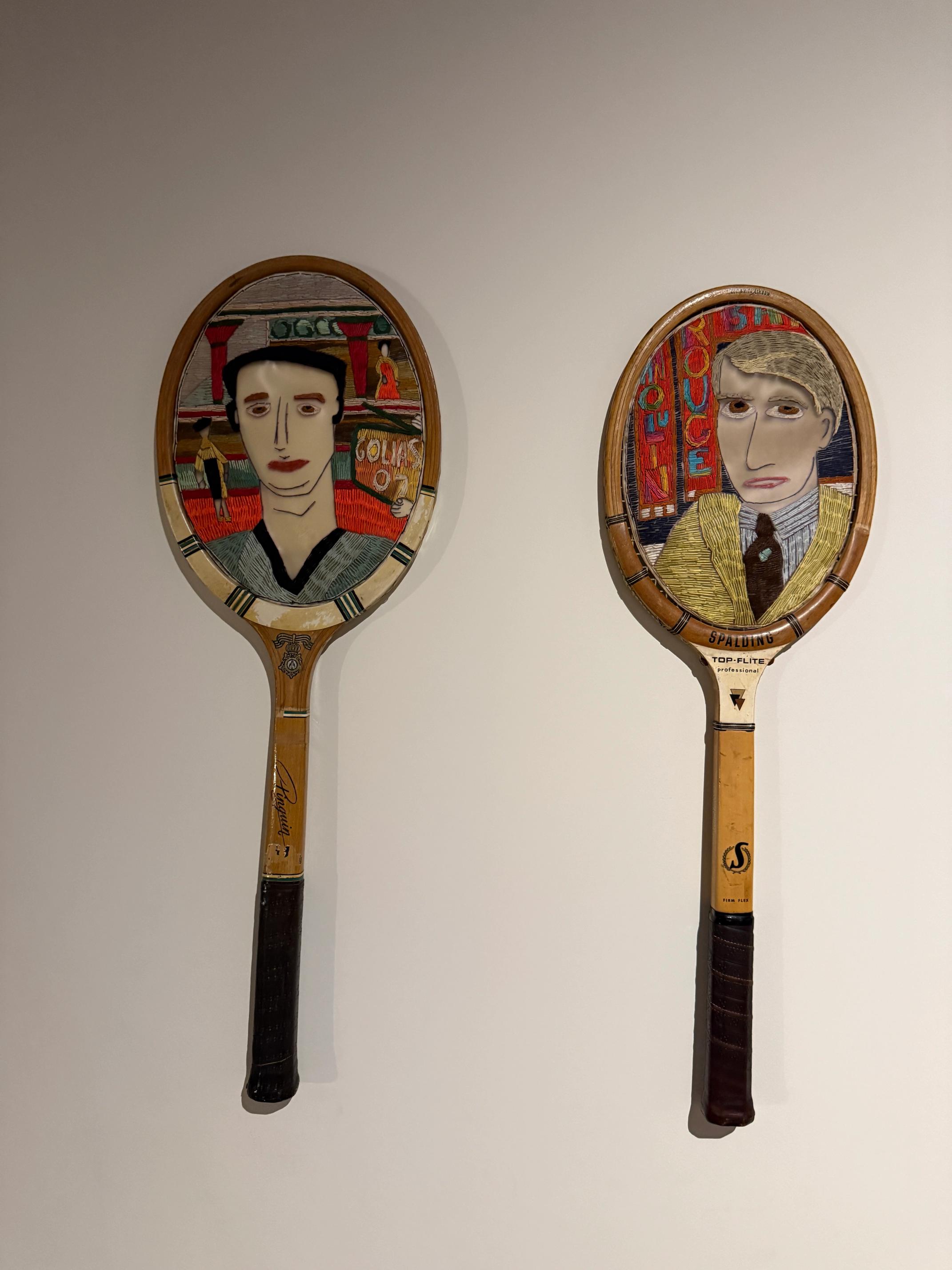 Two vintage wooden tennis rackets, each displaying a painted portrait in its head, hang on a wall.