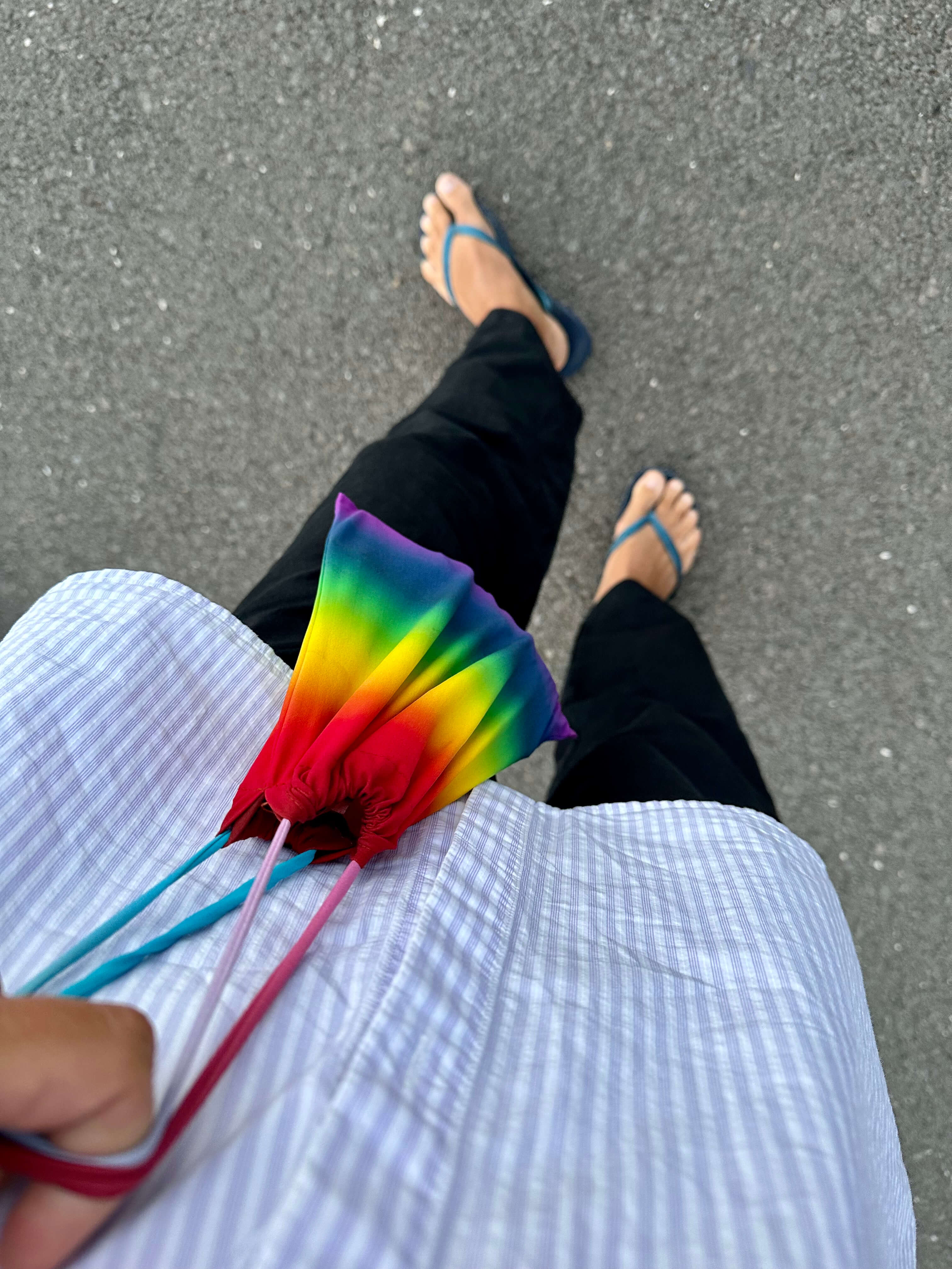 A person wearing a striped shirt, black pants, and blue flip-flops holds a rainbow drawstring bag on asphalt.