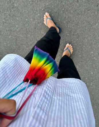 A person wearing a striped shirt, black pants, and blue flip-flops holds a rainbow drawstring bag on asphalt.