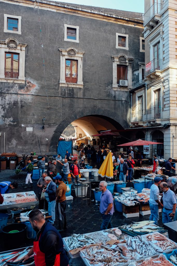 A crowded outdoor fish market in front of a weathered stone building with a large archway, displaying fresh fish.