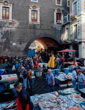 A crowded outdoor fish market in front of a weathered stone building with a large archway, displaying fresh fish.