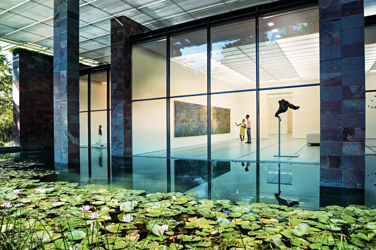 A pond filled with water lilies in the foreground, reflecting a modern art museum with large glass walls. Inside, people look at paintings and sculptures.