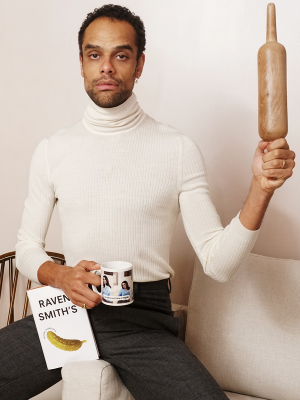 A man in a cream turtleneck holds a wooden rolling pin, a mug with two women pictured, and a book featuring a pickle.
