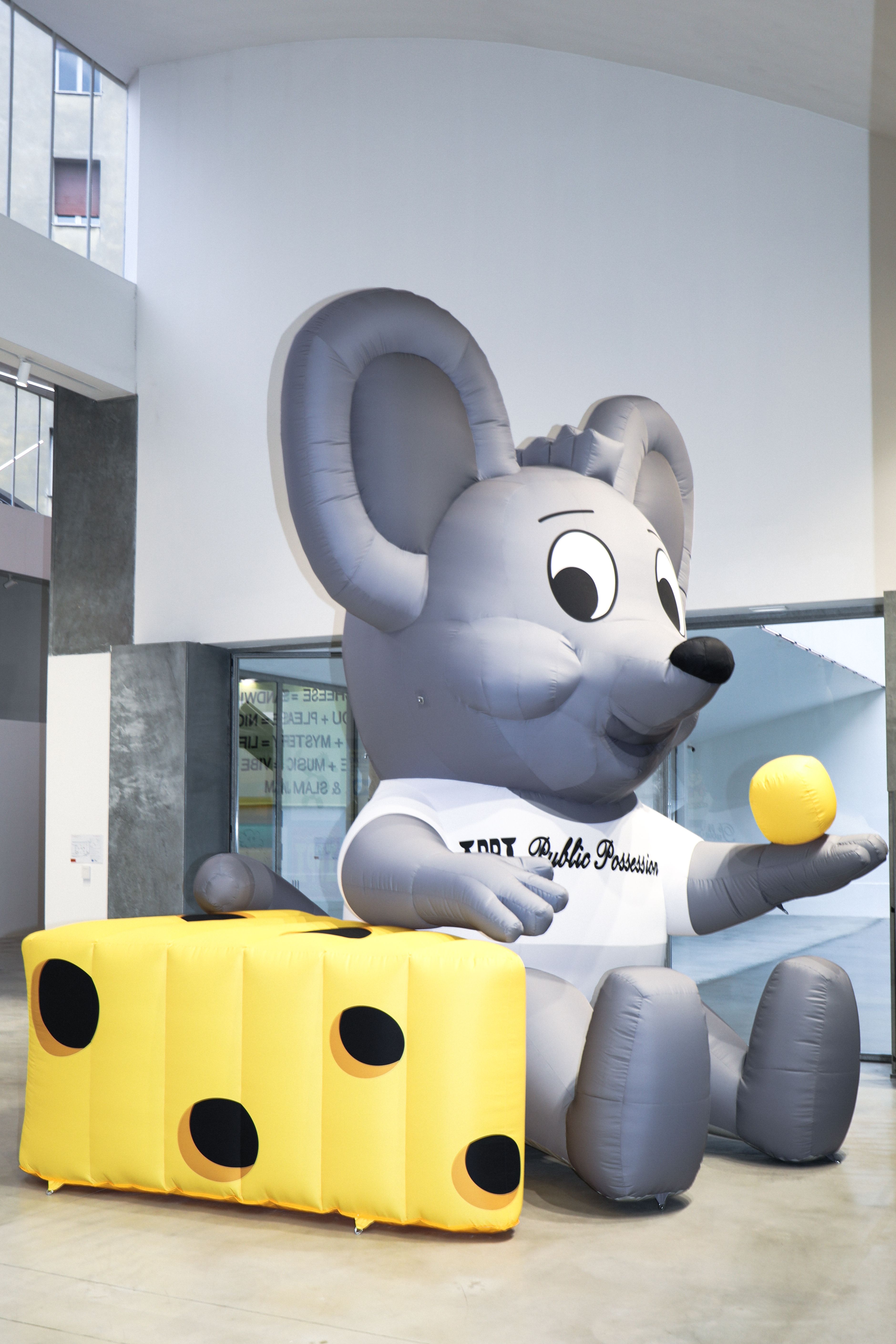 A large inflatable grey mouse mascot wearing a white shirt sits next to an inflatable yellow block of Swiss cheese, holding a yellow ball.