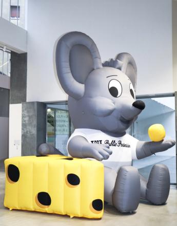 A large inflatable grey mouse mascot wearing a white shirt sits next to an inflatable yellow block of Swiss cheese, holding a yellow ball.