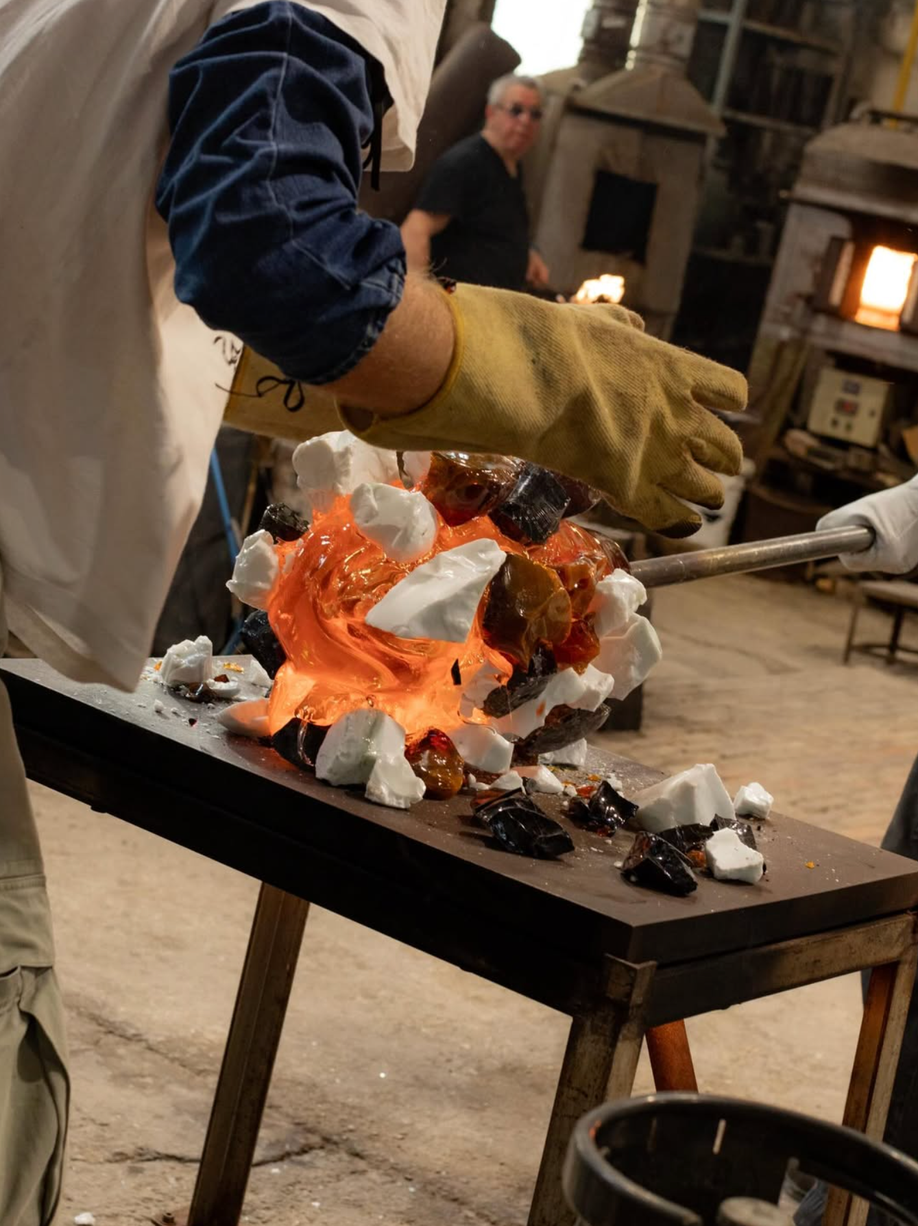 A gloved glassblower works with glowing orange molten glass mixed with white and dark fragments on a table.