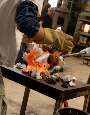 A gloved glassblower works with glowing orange molten glass mixed with white and dark fragments on a table.