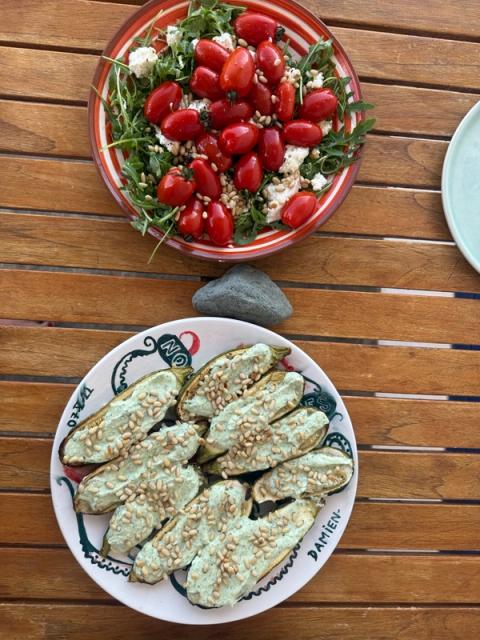 A salad of cherry tomatoes, arugula, feta, and pine nuts, alongside stuffed zucchini with a green sauce and pine nuts on a wooden table.