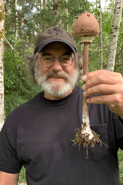 A bearded man in glasses and a cap with a mushroom emblem holds a large, scaly brown mushroom in a forest.