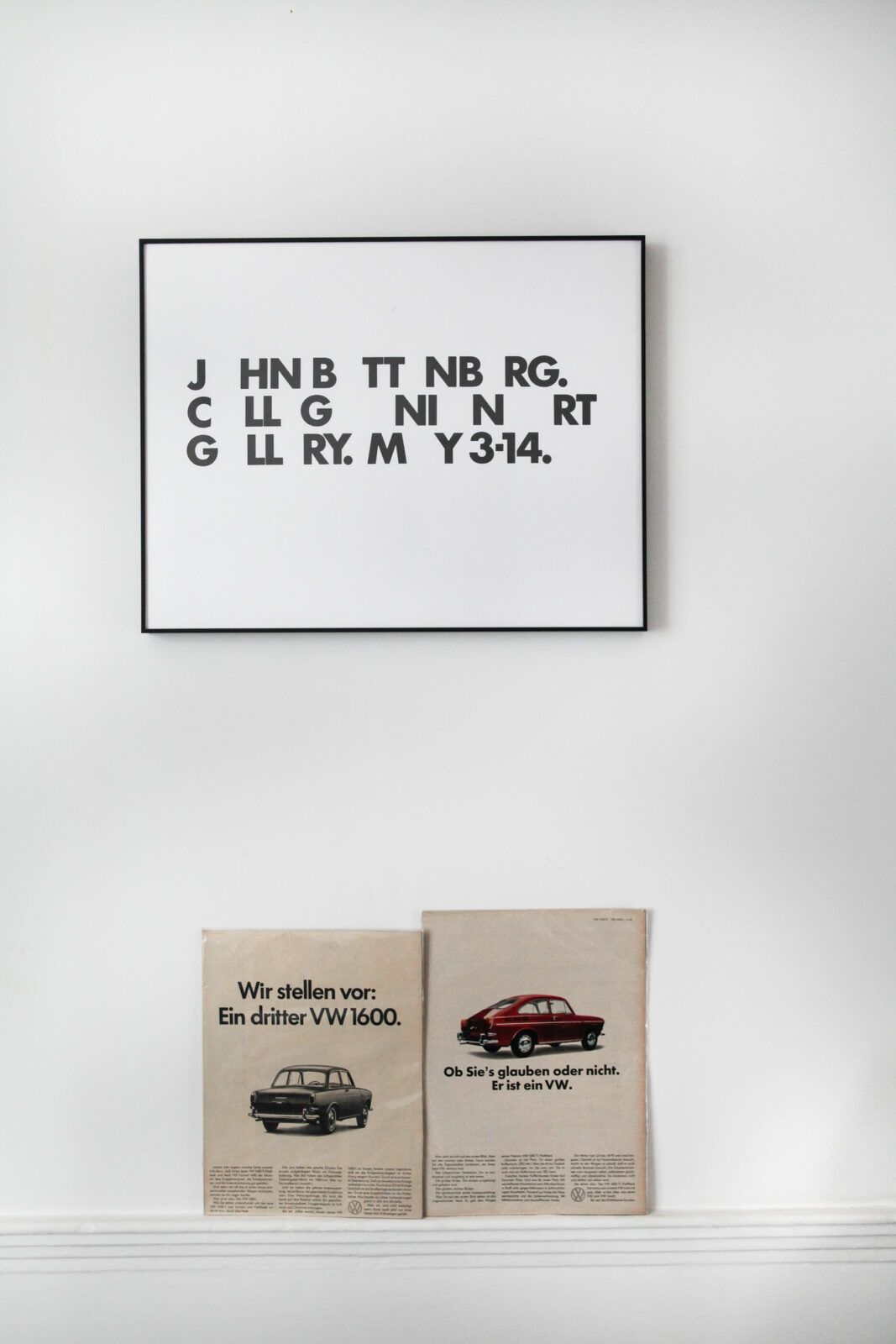 A framed poster with stylized black text hangs above two vintage Volkswagen car advertisements on a white shelf.
