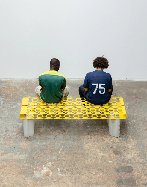 Two people sit on a yellow honeycomb-patterned bench, seen from behind. One wears a navy shirt with "75" on the back.