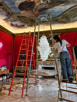A woman on a ladder adjusts a large chandelier made of irregular white glass pieces in a grand room with red walls and a painted ceiling.