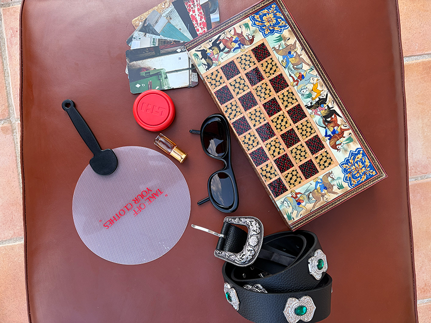 A decorative wooden chessboard, black sunglasses, a black belt with ornate silver and green buckles, and a translucent fan with 'TAKE OFF YOUR CLOTHES' in red text, laid on a brown surface.