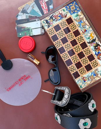 A decorative wooden chessboard, black sunglasses, a black belt with ornate silver and green buckles, and a translucent fan with 'TAKE OFF YOUR CLOTHES' in red text, laid on a brown surface.