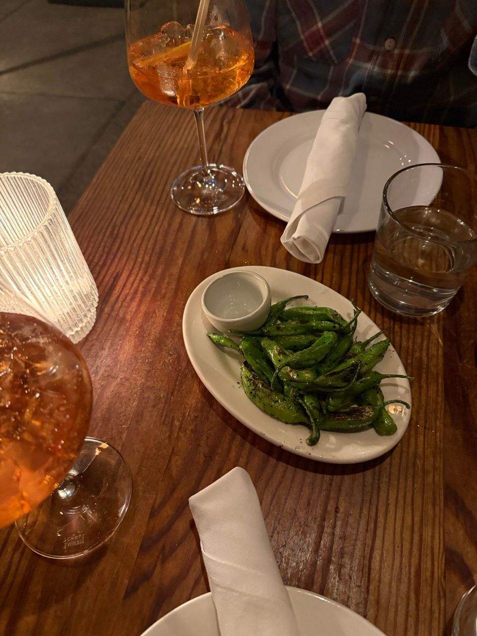 A wooden table set with two orange drinks, a plate of grilled green peppers, a glass of water, and white napkins.