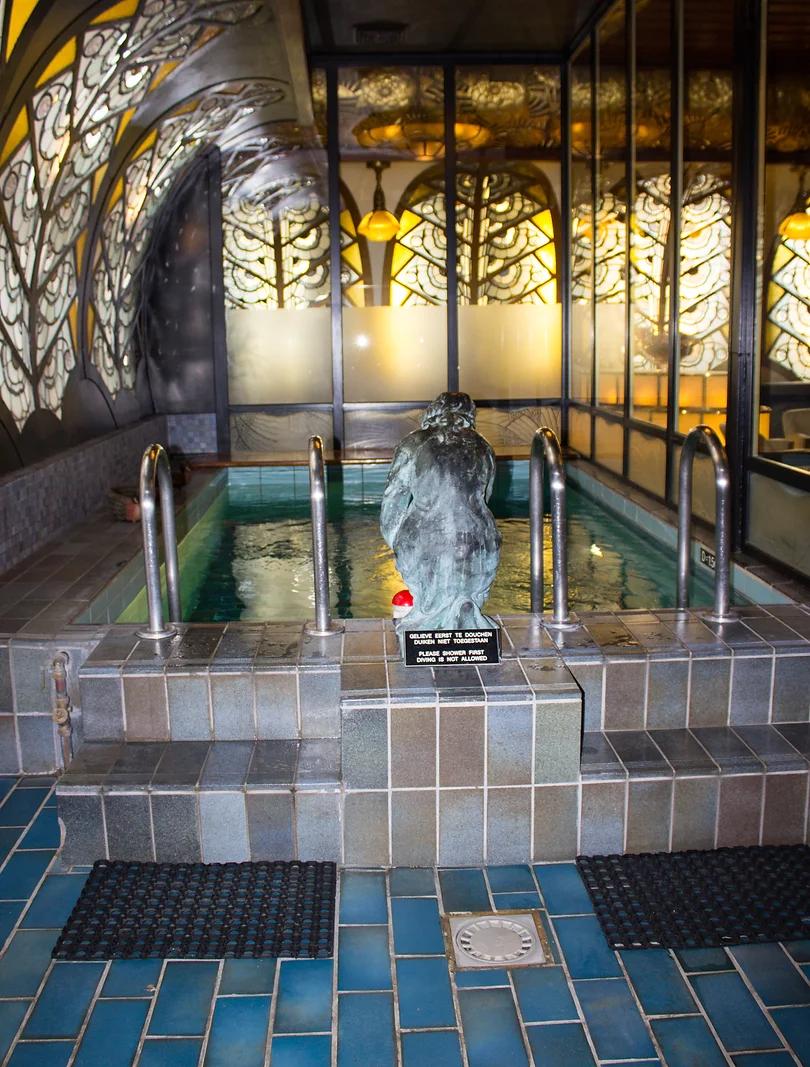 An indoor pool with a statue, surrounded by ornate, glowing stained glass panels and tiled surfaces.