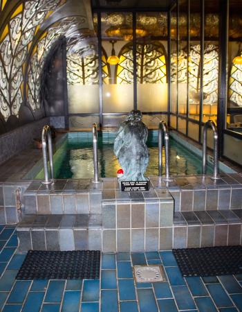 An indoor pool with a statue, surrounded by ornate, glowing stained glass panels and tiled surfaces.