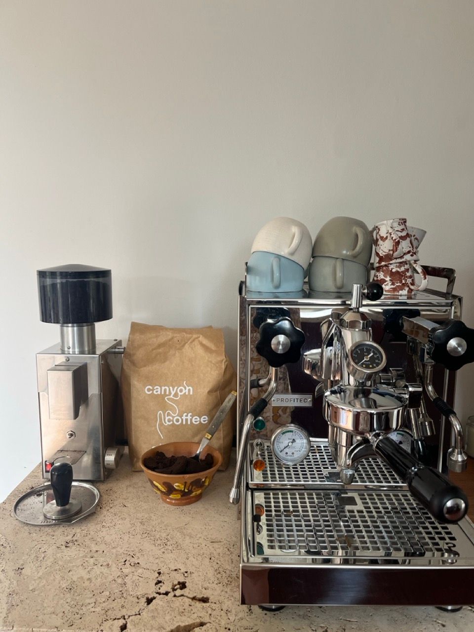 A coffee setup with a stainless steel grinder, a bag of Canyon Coffee, a Profitec espresso machine, and stacked cups on a travertine counter.
