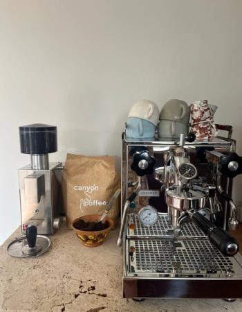 A coffee setup with a stainless steel grinder, a bag of Canyon Coffee, a Profitec espresso machine, and stacked cups on a travertine counter.